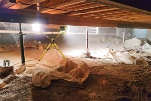 Crawl space part-way through encapsulation, with heavy vapor barrier laid out across the dirt floor, adjustable support columns in place, and a work light on a tripod lighting the joists overhead
